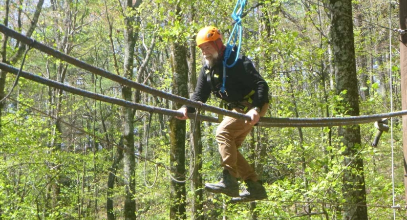 A person wearing safety gear is attached to ropes as they navigate a ropes course in a wooded area. 